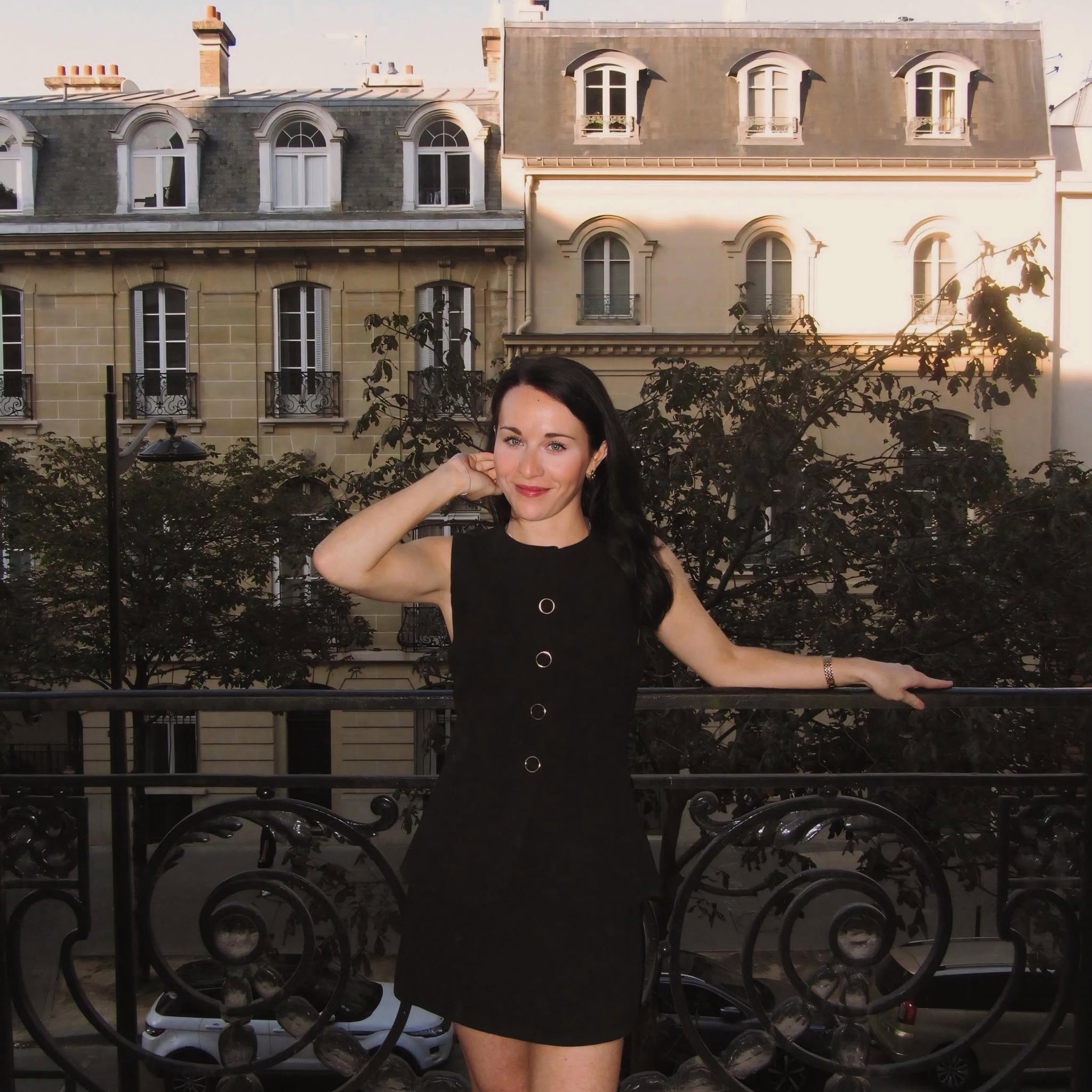 Woman with dark hair in a black dress standing on a balcony with Parisian buildings in background.