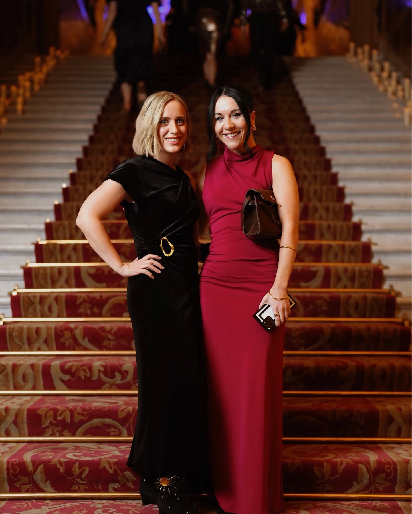Two women in elegant dresses standing on a staircase, smiling, with a blurred background of people and warm lighting.