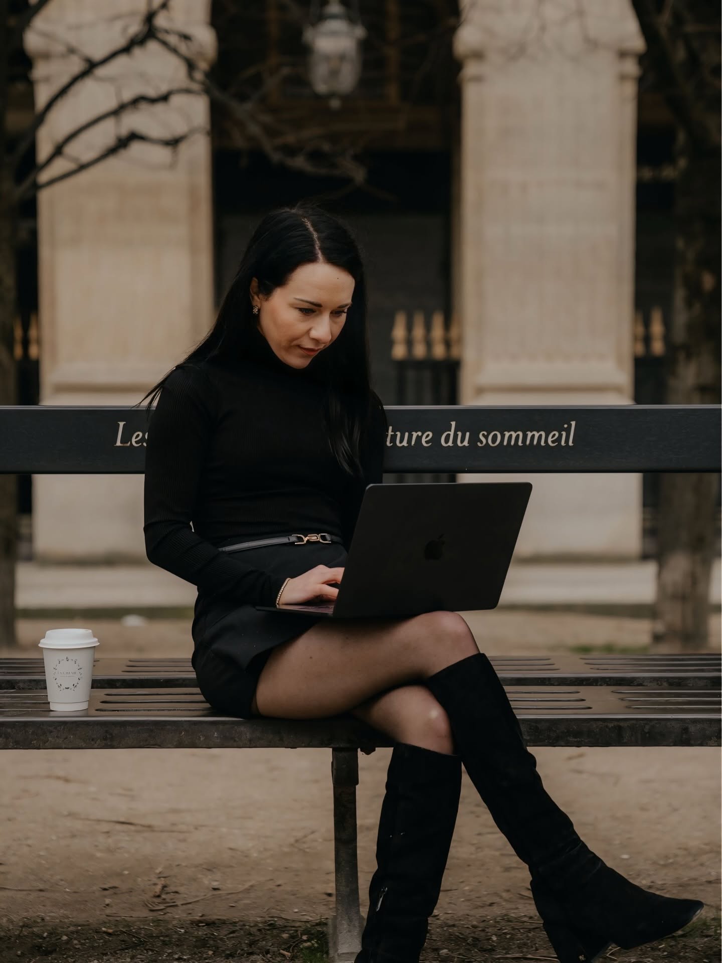 Woman sitting on a bench using a laptop outdoors, with architectural columns in the background.
