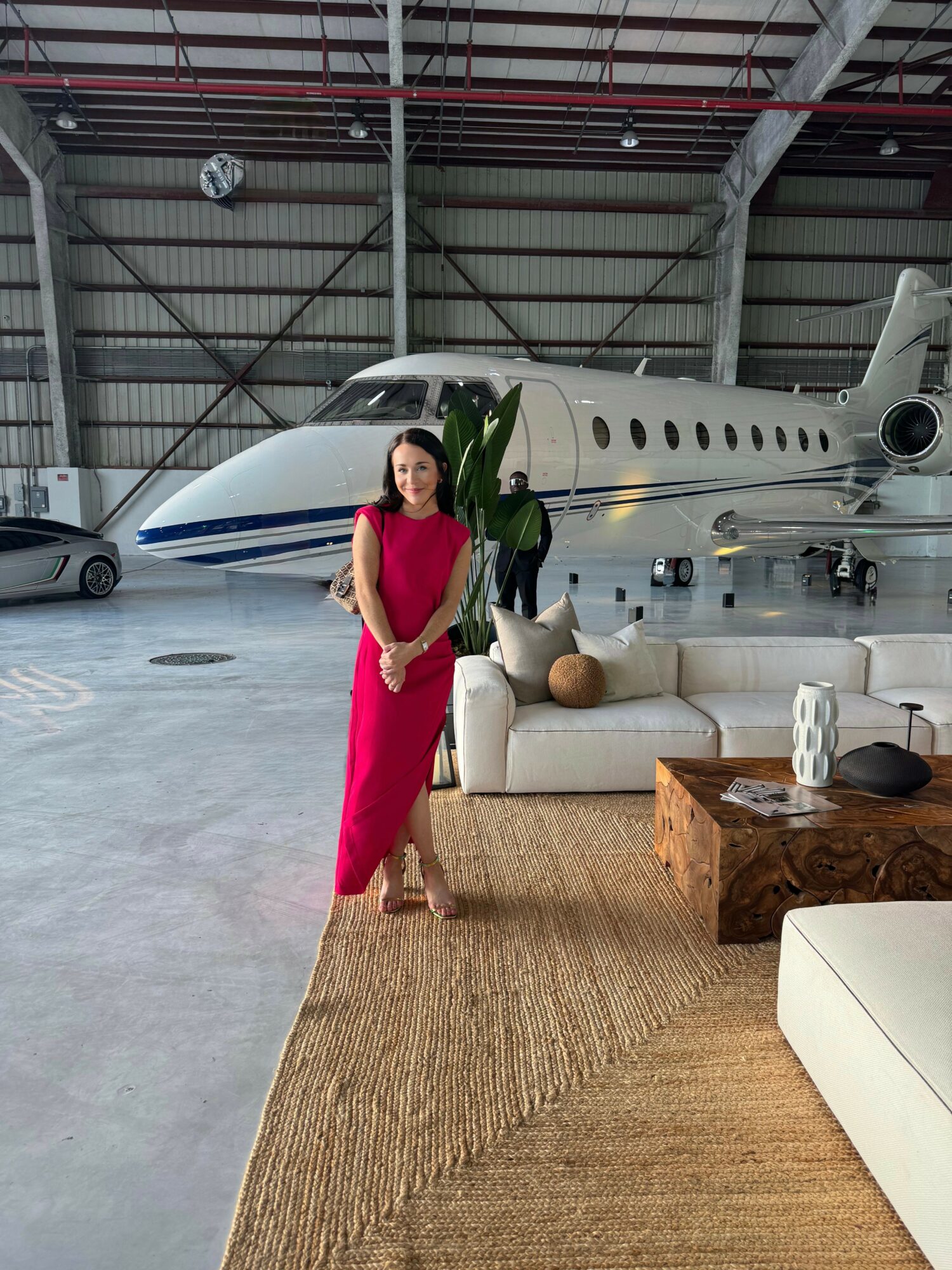 Woman in pink dress standing inside hangar with private jet, sofa, and wooden table.