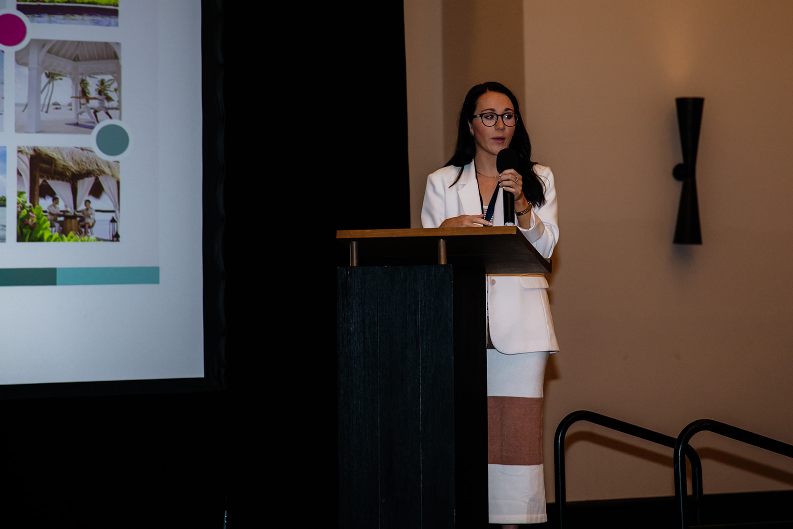Woman in white blazer and glasses standing at a podium giving a presentation, with a projection screen on the left.