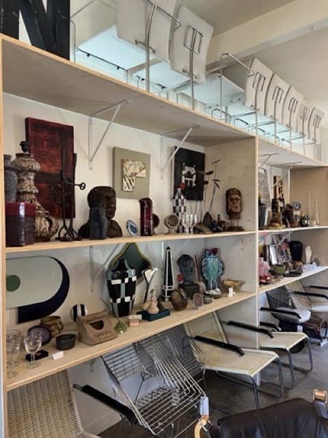 Shelves with various decorative items, sculptures, and vases, with chairs lined up above and below, in a retail or gallery setting.