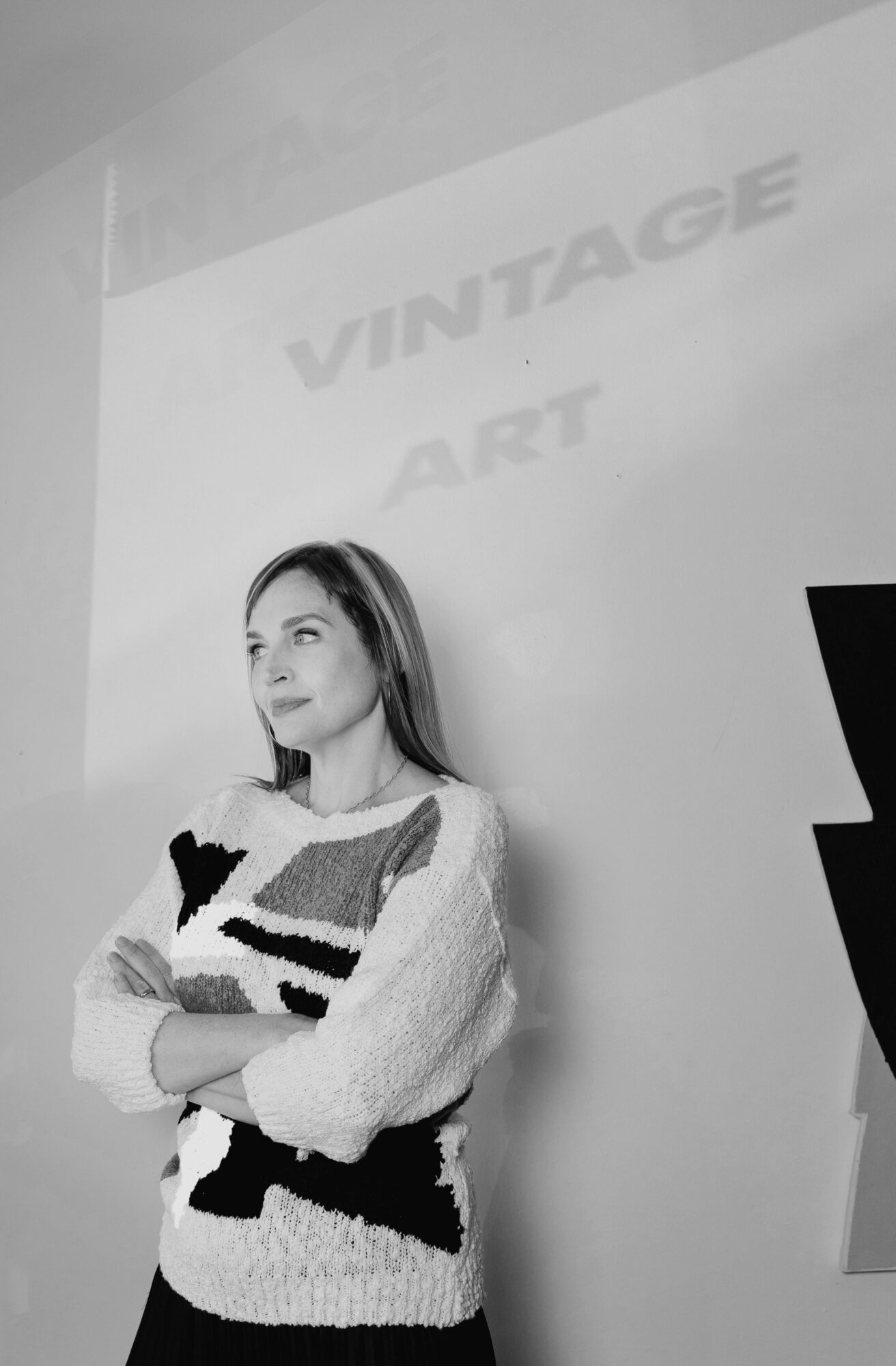 Young woman with long hair standing with arms crossed, in front of a wall with text, in black and white.