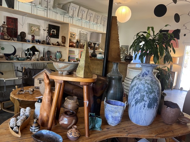 Display of vases, plants, and decorative objects on a wooden table in a room with shelves and artwork.