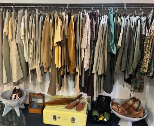 Clothes hanging on a rack, shoes on a yellow suitcase, and various items on the floor, including bags and boxes.