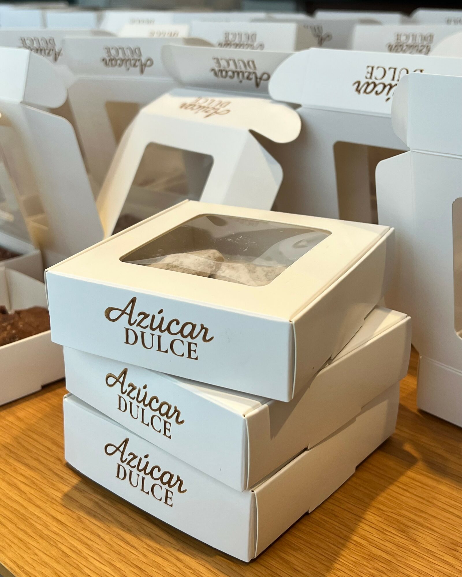 Three stacked white boxes with transparent windows, labeled 'Azúcar Dulce,' on a wooden surface, with more boxes in the background.