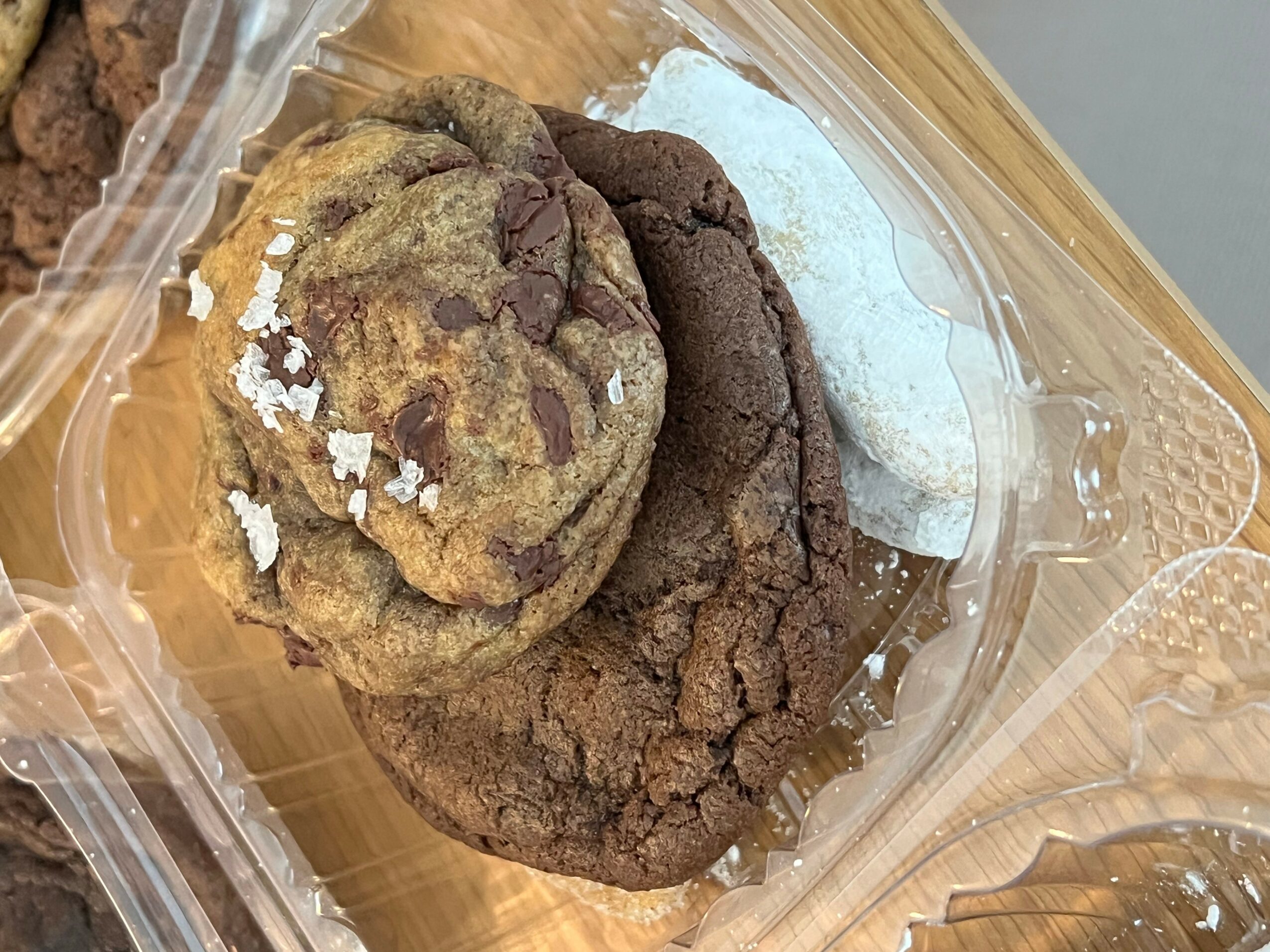 Three cookies in a clear plastic container, one with chocolate chips, one plain, and one with white sugar, on a wooden surface.