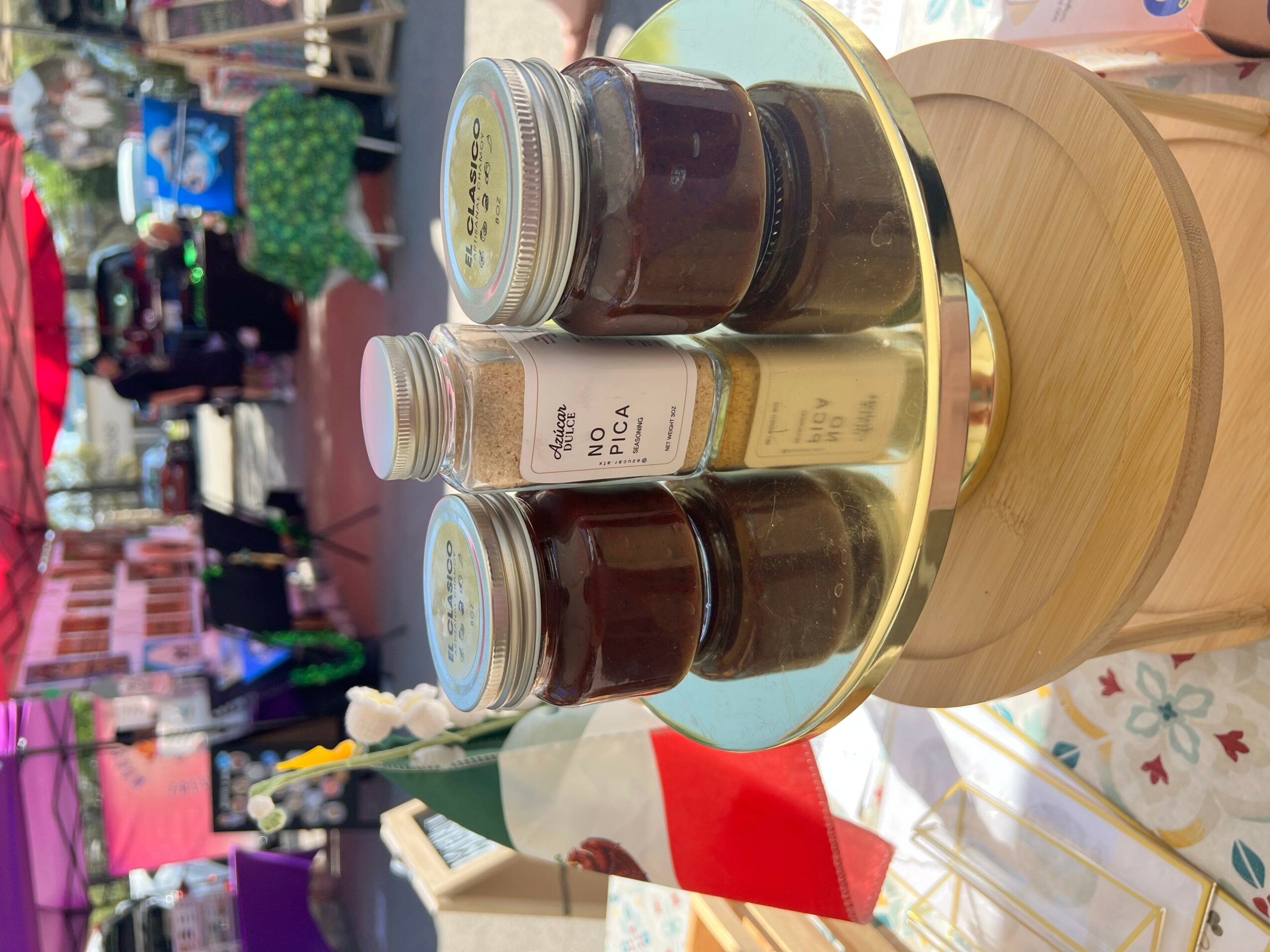 Round wooden stand with jars of jam and honey on a table at an outdoor market, colorful stalls in background.