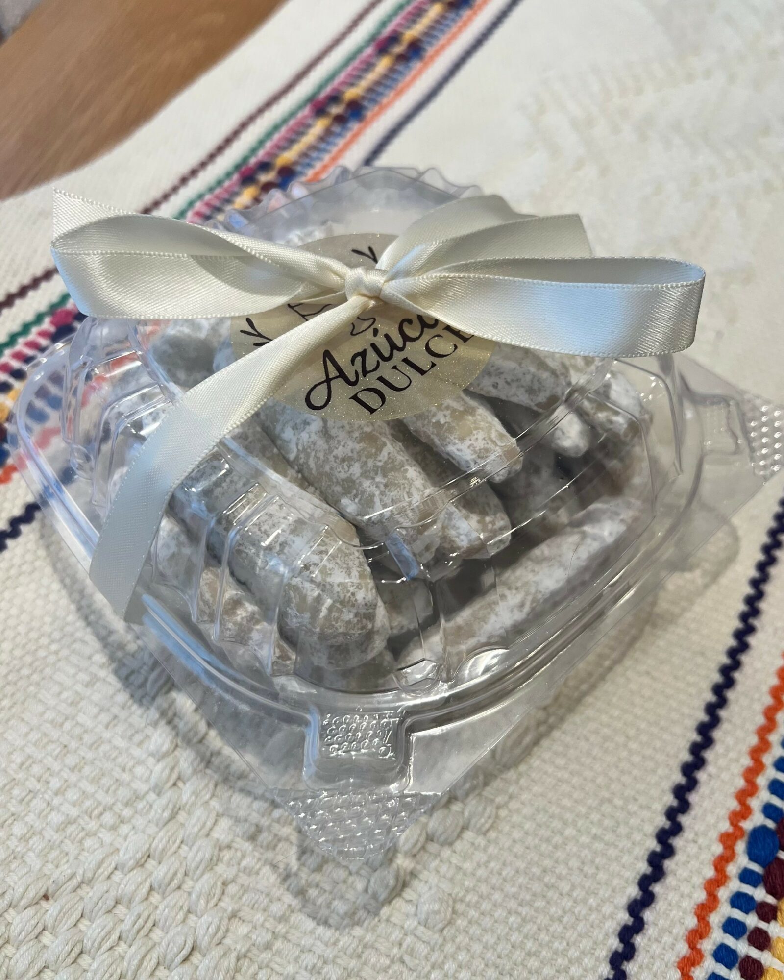Clear plastic container with wrapped candies tied with a white ribbon, placed on a white cloth with colorful embroidery.