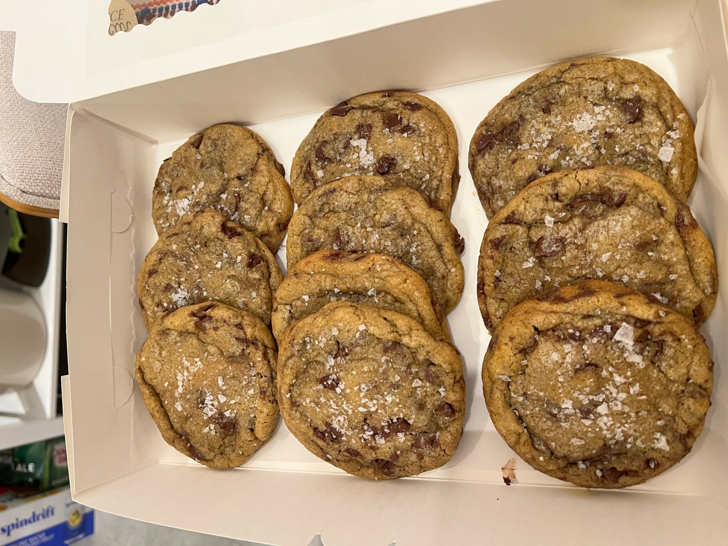 Nine chocolate chip cookies in a white box, some with visible chocolate chips, arranged in two rows.