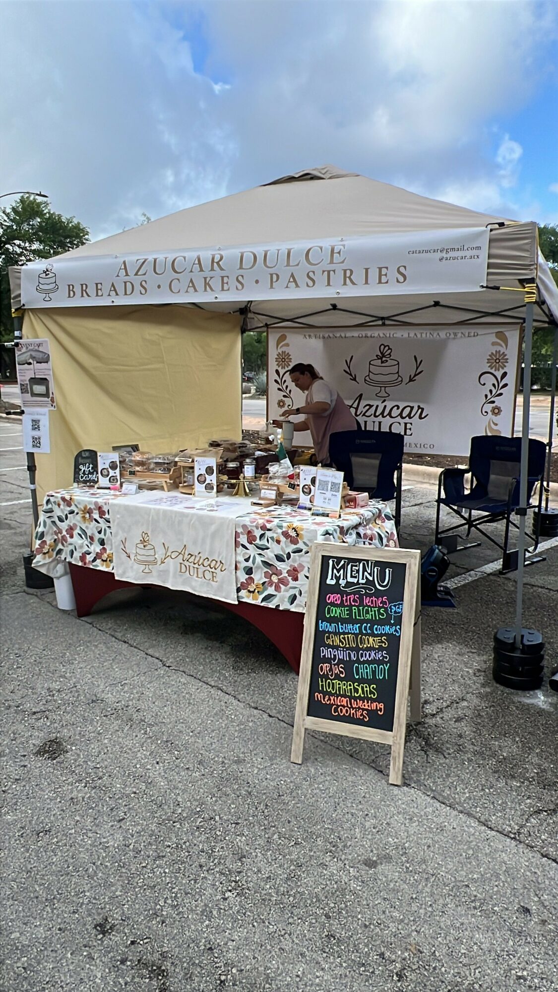 Market stall with a white canopy selling baked goods, with a chalkboard sign outside, on a sidewalk.