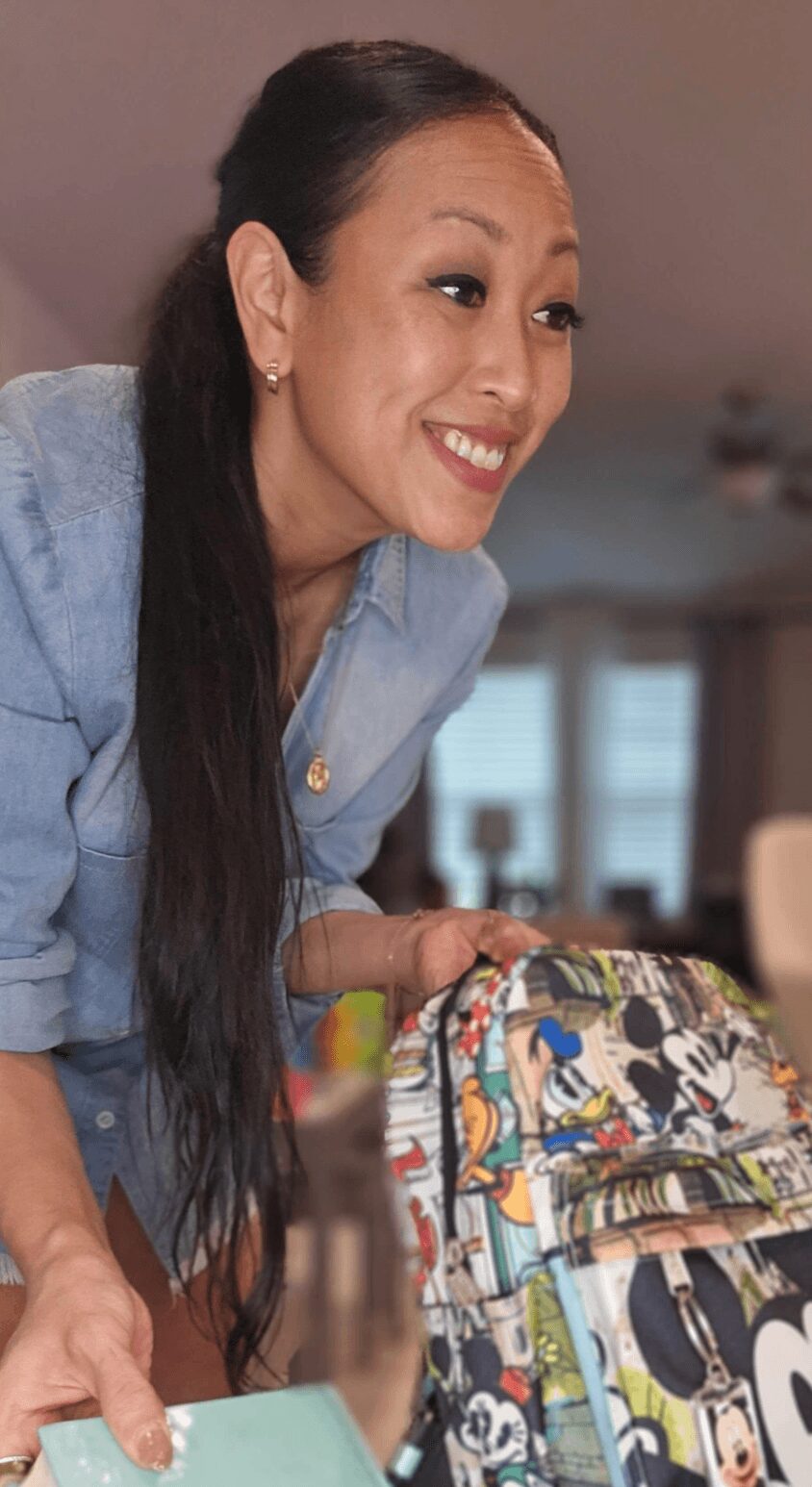 Smiling woman with long dark hair leaning forward, wearing a light blue shirt, in a room with windows and furniture