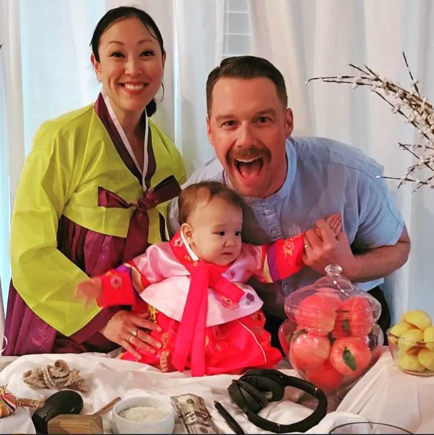 Family of three smiling at a table with fruit and decorations, woman in traditional dress, man and baby girl in colorful clothing