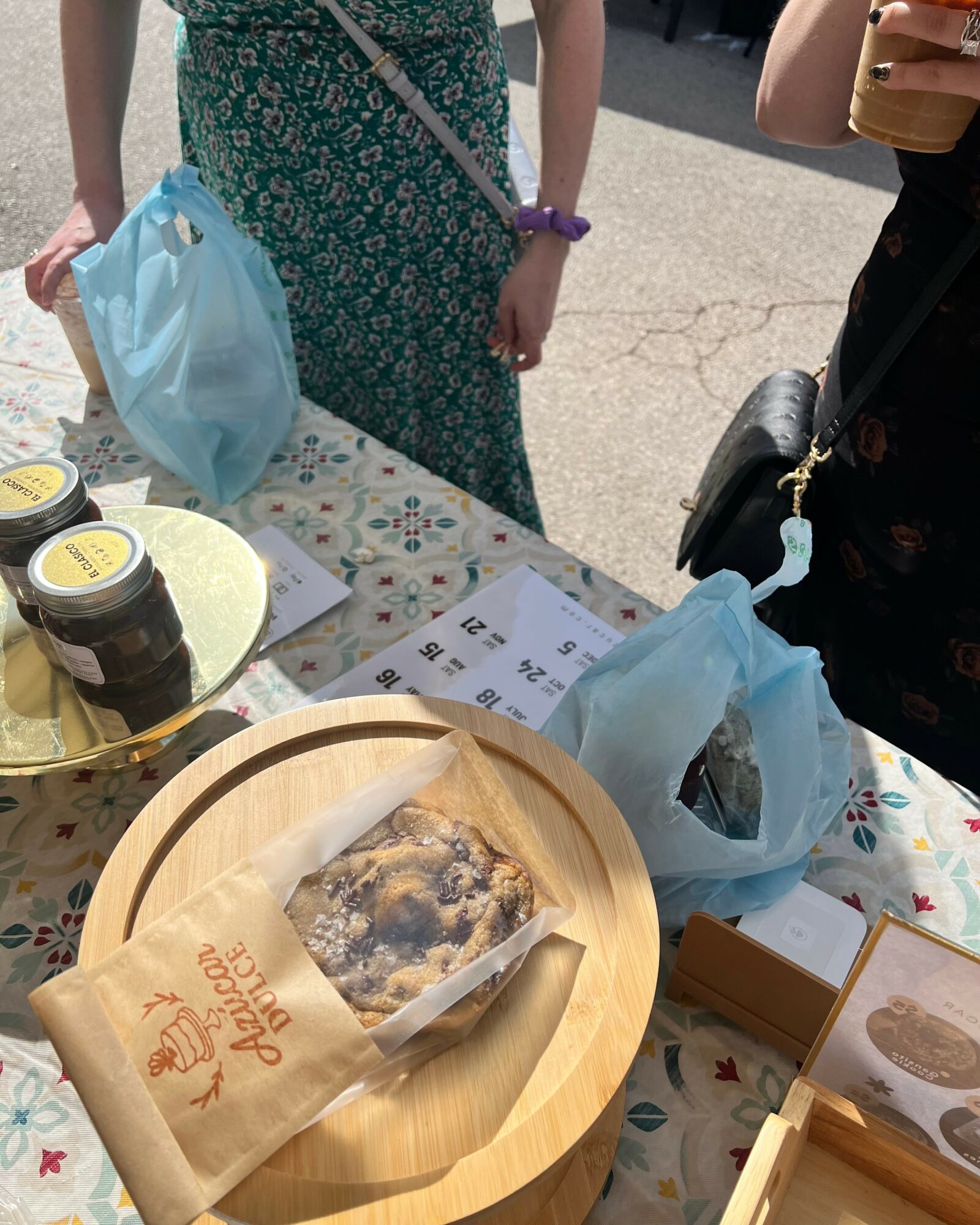 People standing around a table with food jars, a wooden tray with a packaged dessert, and blue bags outdoors.
