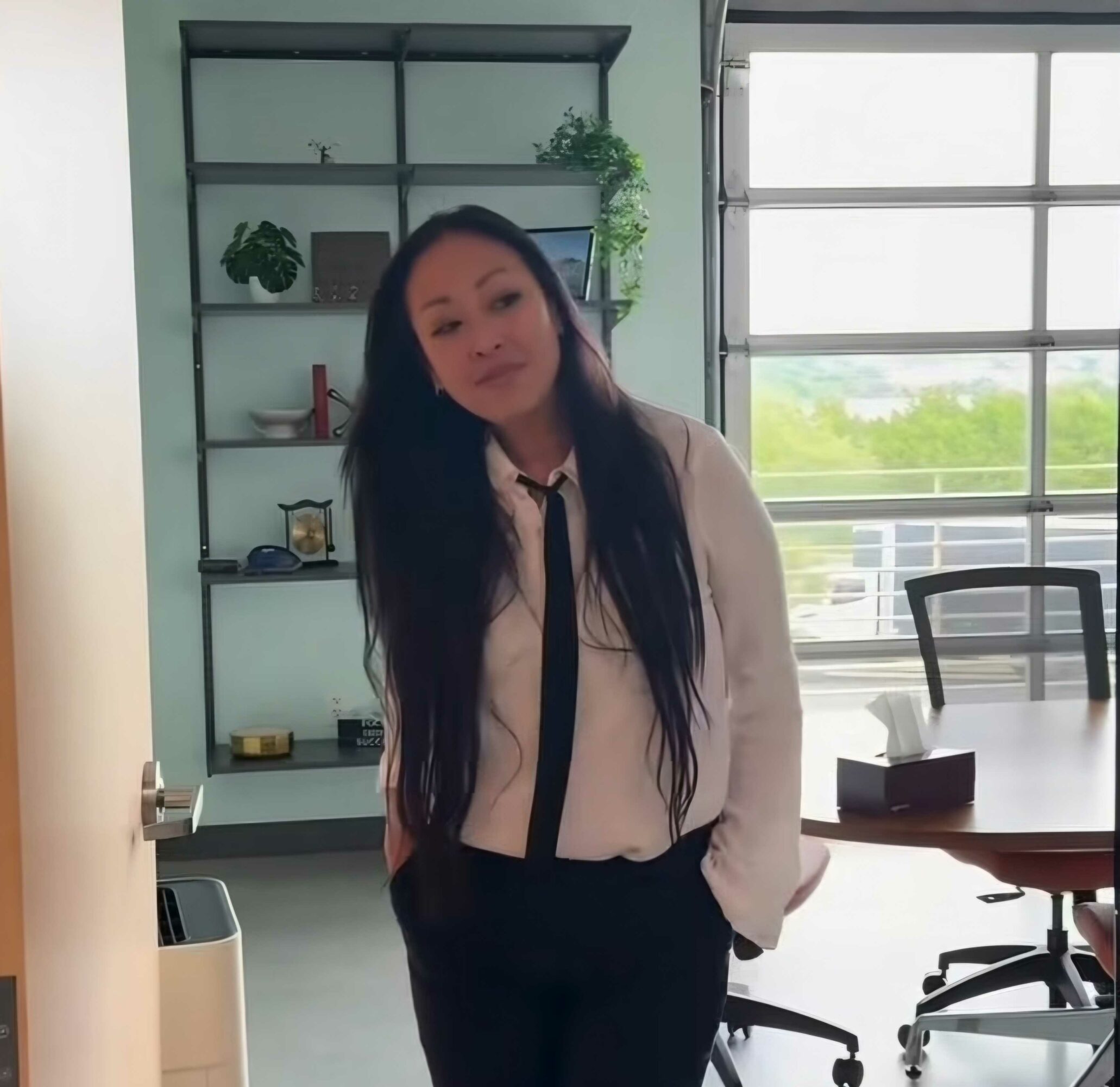 Young woman with long dark hair in a white shirt and black pants stands in an office, with a bookshelf and window behind her.