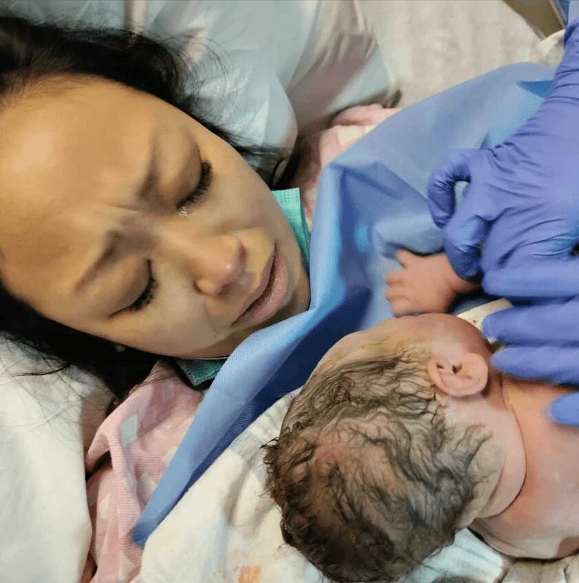 Woman lying in hospital bed holding newborn baby with medical staff's gloved hand touching baby's head.