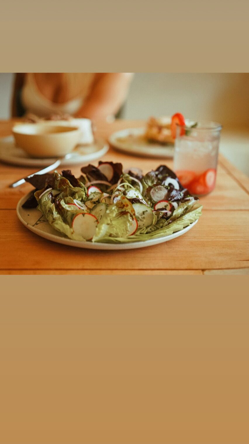 Plate of salad with vegetables and dressing on a wooden table, blurred person and drink in background.