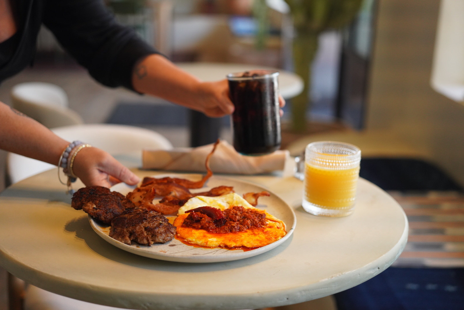 Person serving a plate with fried eggs, bacon, and hash browns on a white table, with a glass of orange juice and a dark beverage.