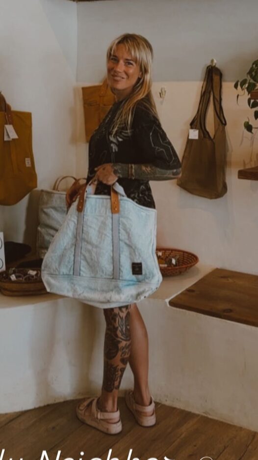 Woman with blonde hair holding a large light blue bag, standing indoors near a wall with hanging bags.