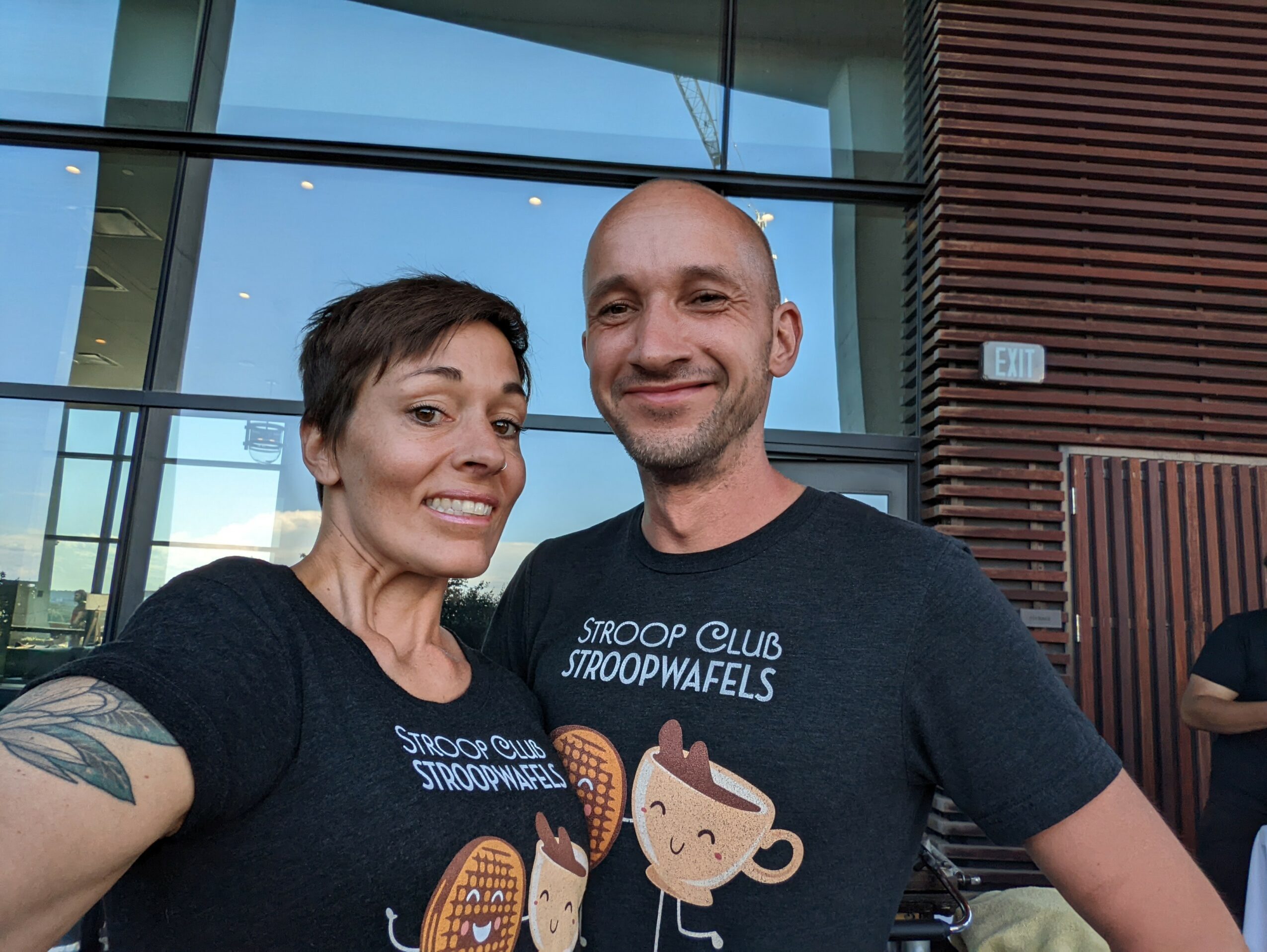 Two smiling people wearing matching black T-shirts with cartoon coffee cups, standing outside a modern building.