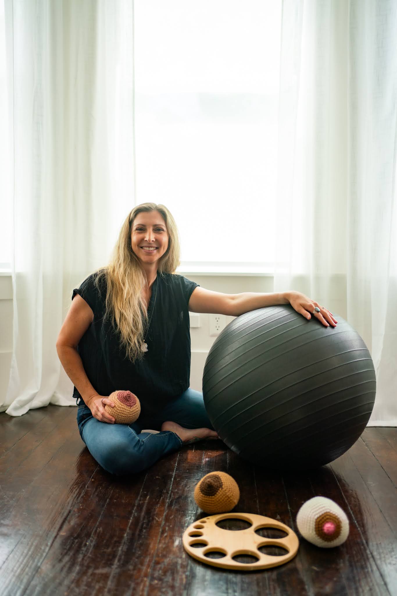 Woman sitting on floor with exercise ball, smiling, in a room with large window and white curtains.