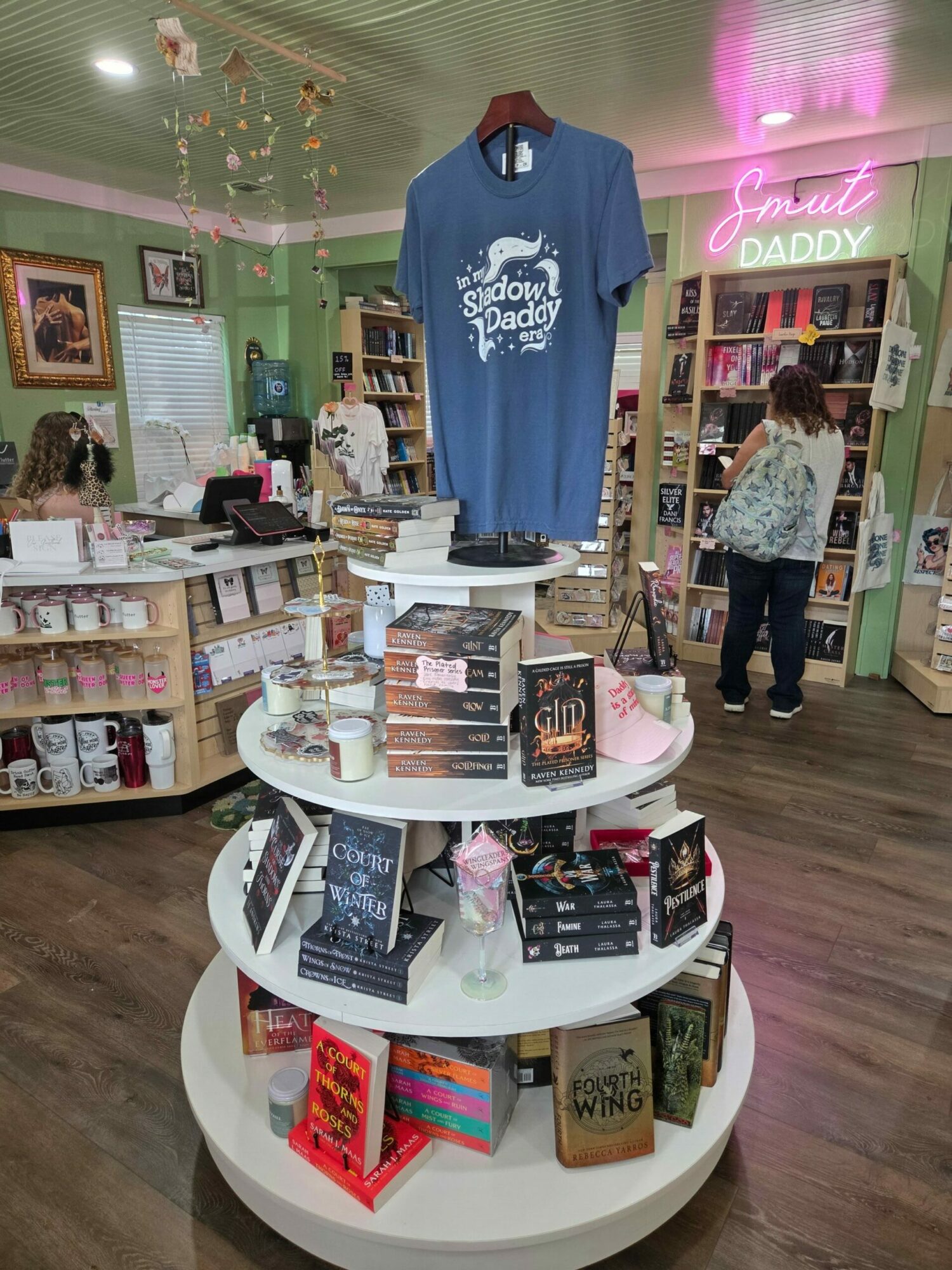 Display table with books, a blue T-shirt, and decorative items inside a bookstore, with shelves and a person in the background.