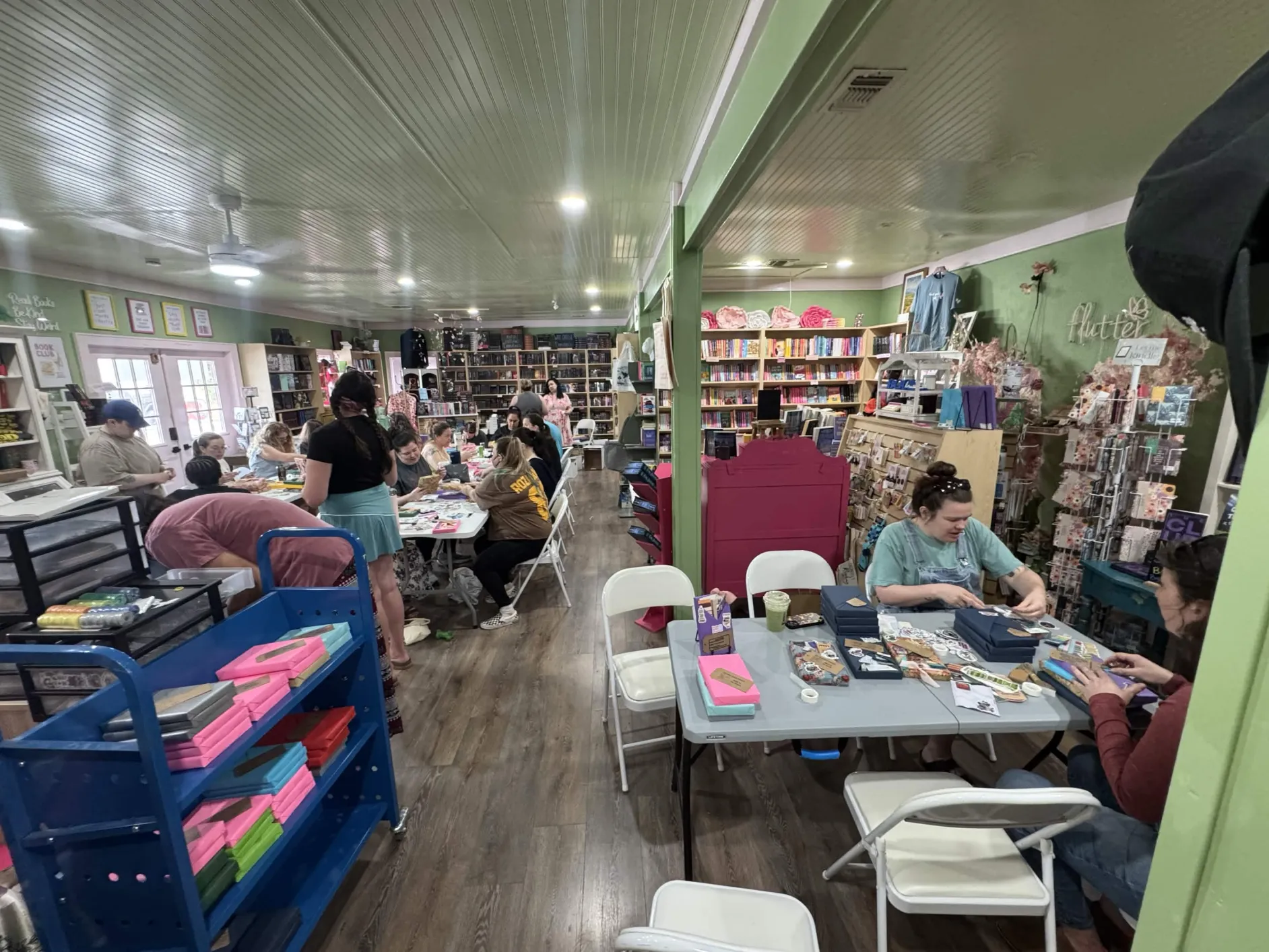People working at tables in a store with shelves of books and supplies, bright lighting, and a green ceiling.