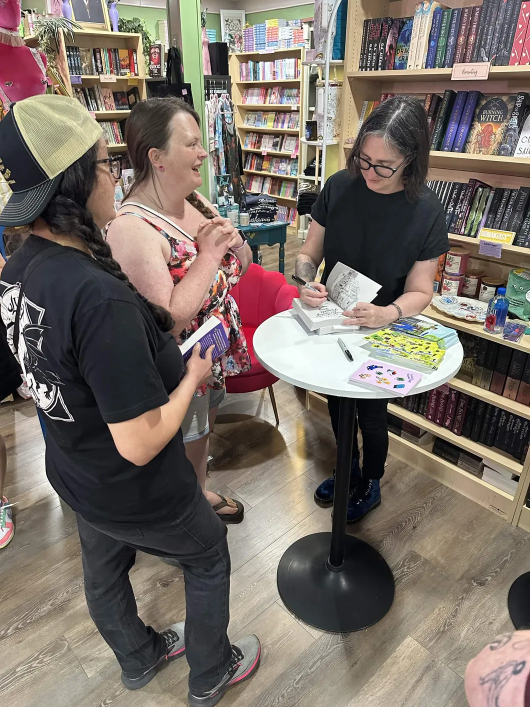 Three women in a bookstore, two holding books and one signing a book at a small round table.