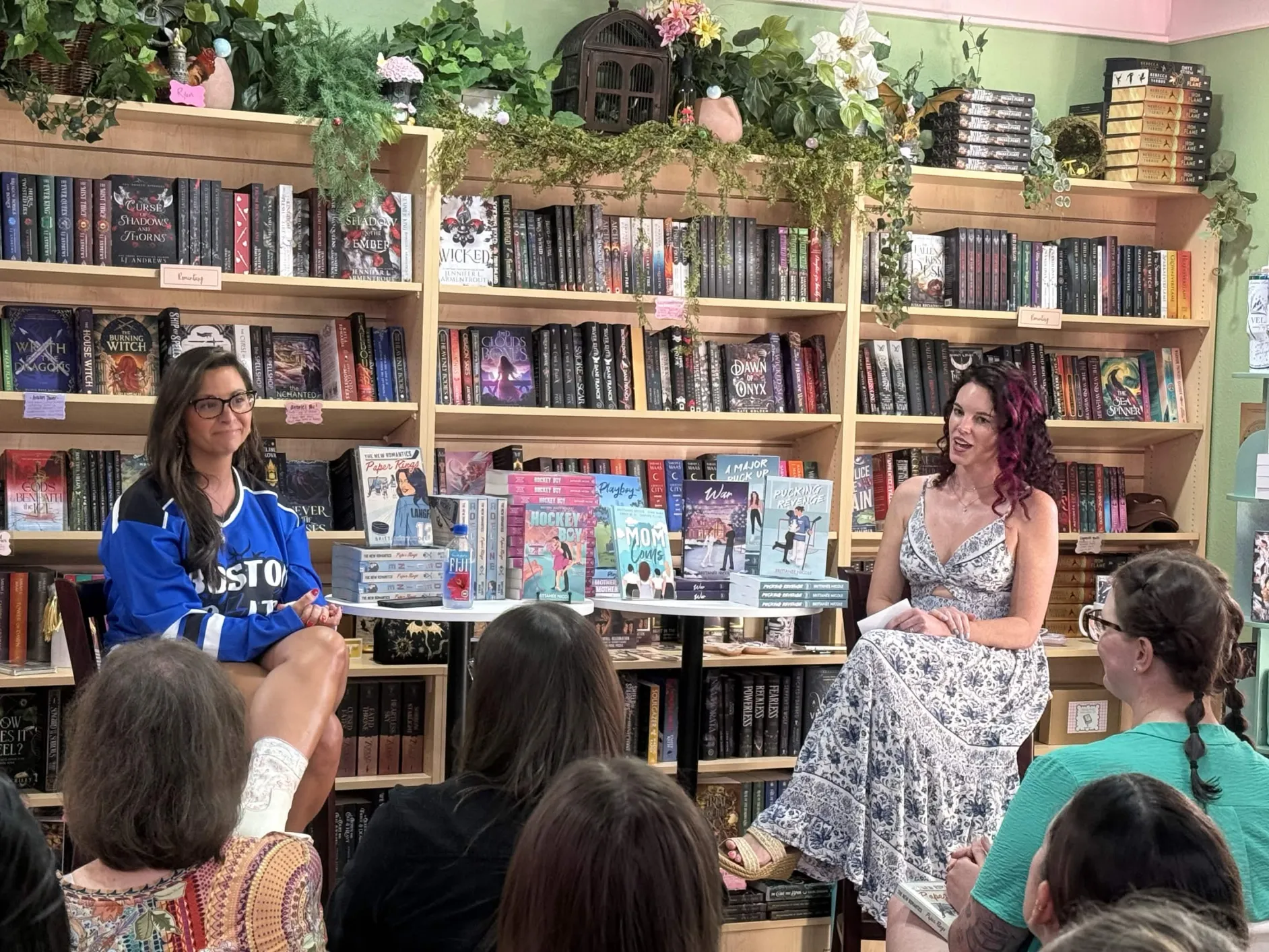 Two women sit in front of a bookshelf filled with books and plants, speaking to an audience in a library.
