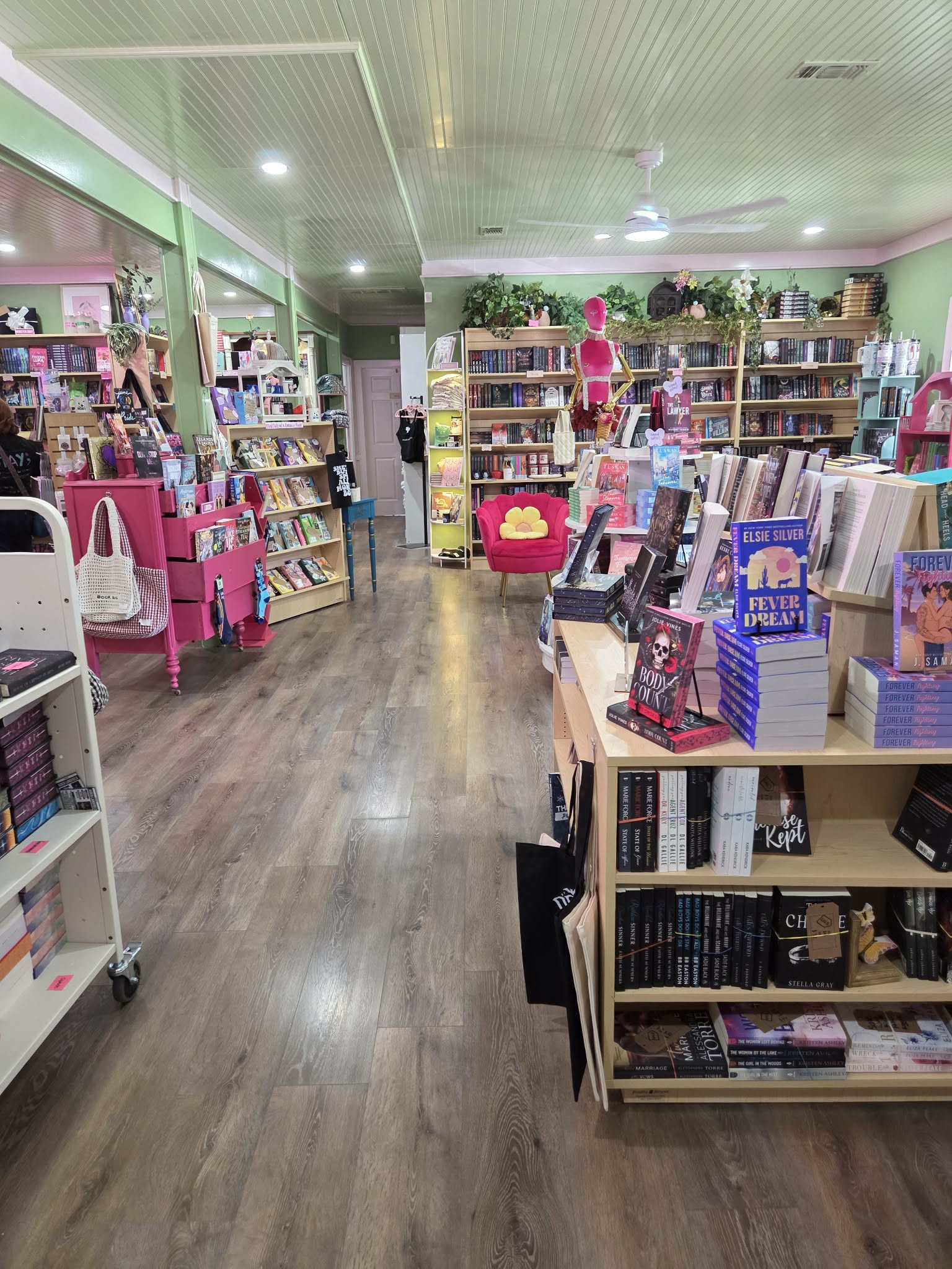 Bookshelves filled with books and items in a brightly lit store with wooden flooring and pink accents.