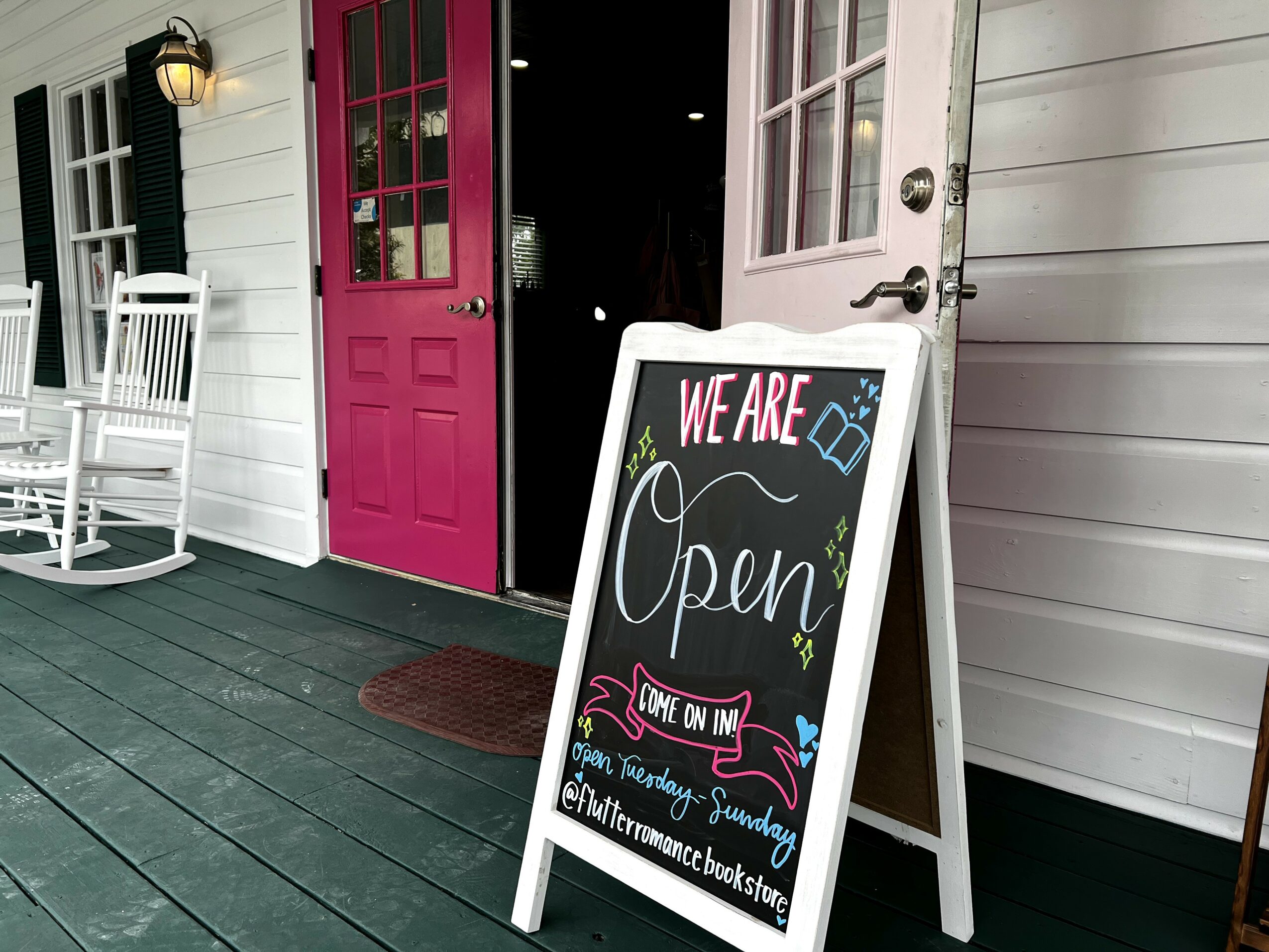 A chalkboard sign outside a building with a pink door, displaying opening hours and message, on a porch with white chairs.
