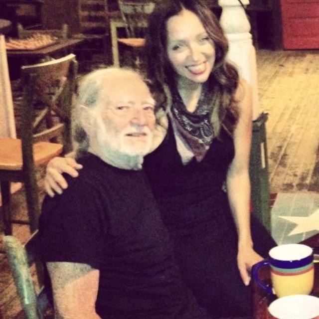 An elderly man with white hair and beard, and a young woman with long dark hair, smiling together indoors.