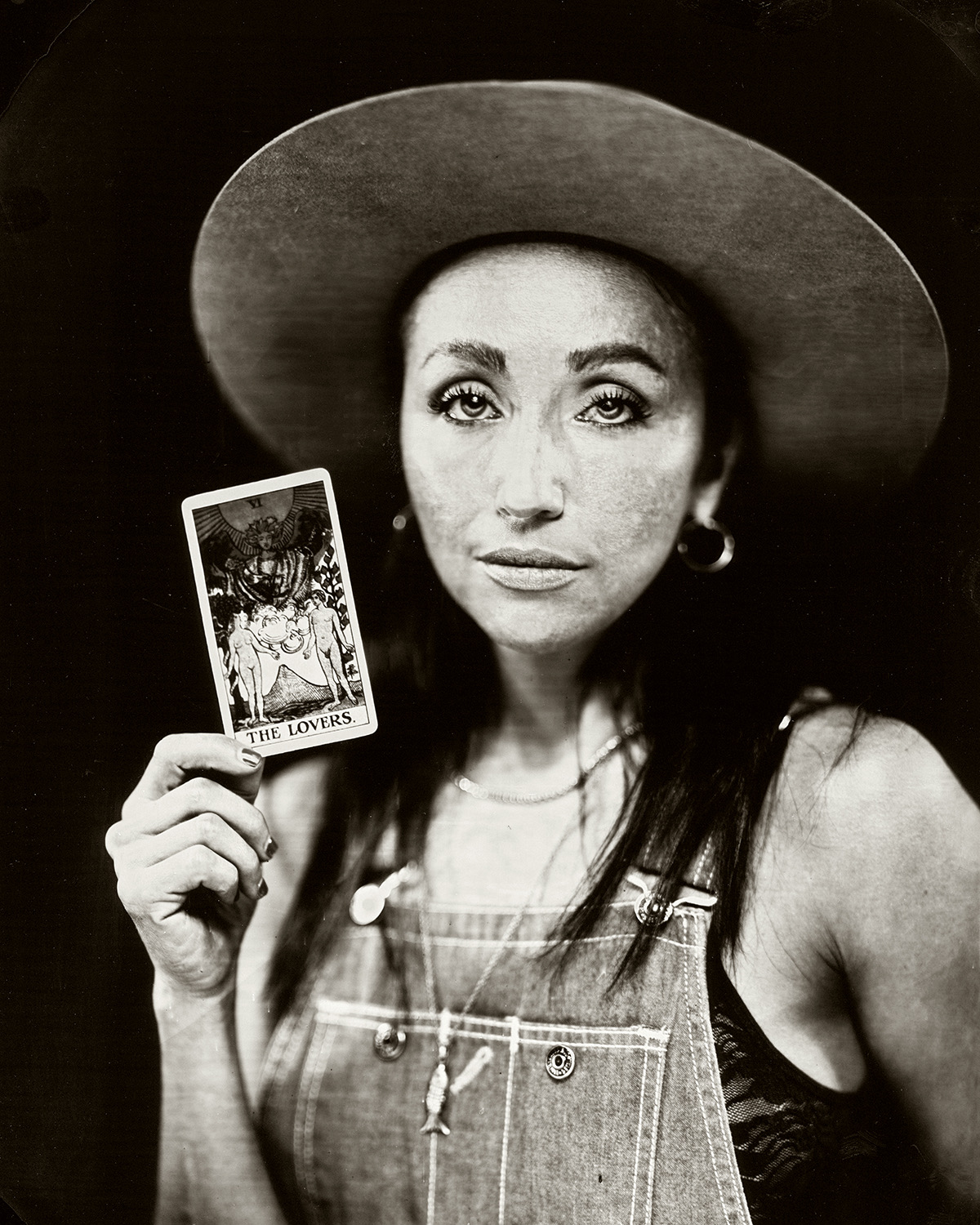 Woman wearing a wide-brimmed hat holding a tarot card, looking directly at the camera, in black and white.