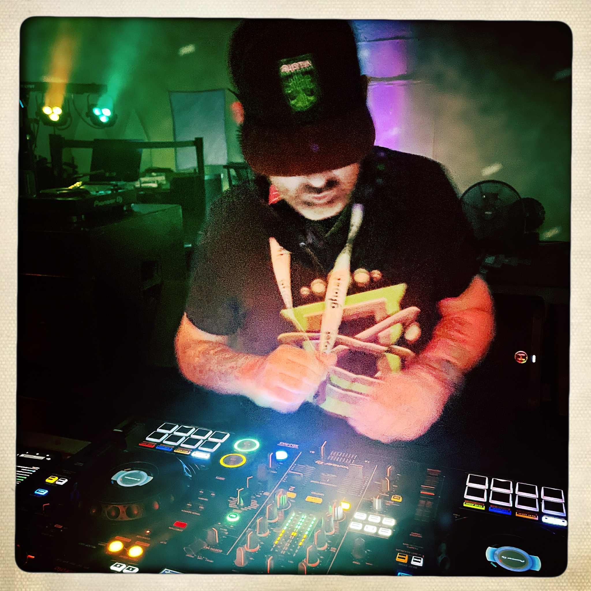 Person wearing a cap and headphones, operating DJ equipment in a dimly lit club with colorful lights.