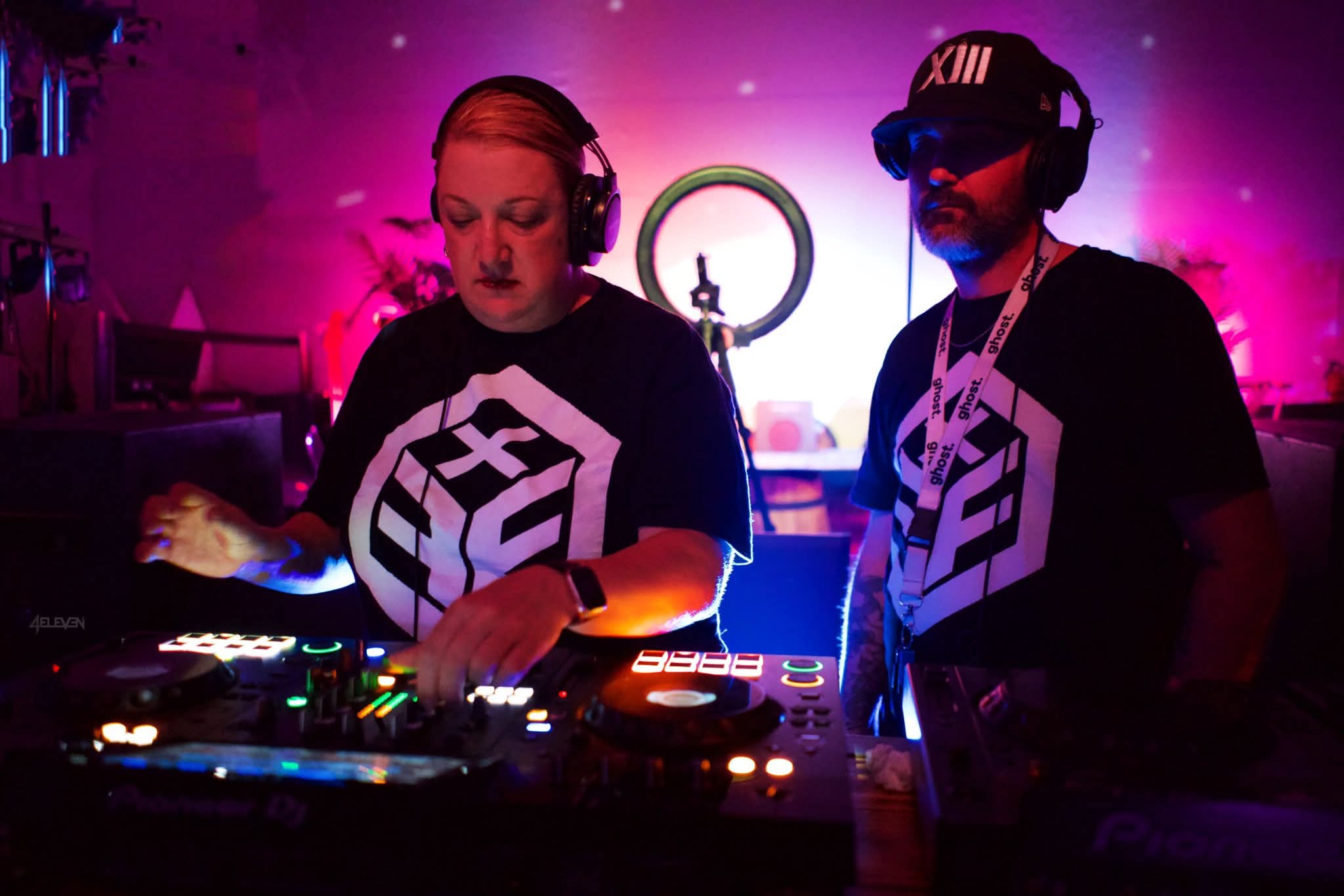 Two DJs wearing headphones operate a mixing console in a colorful, illuminated setting with pink and purple lighting.