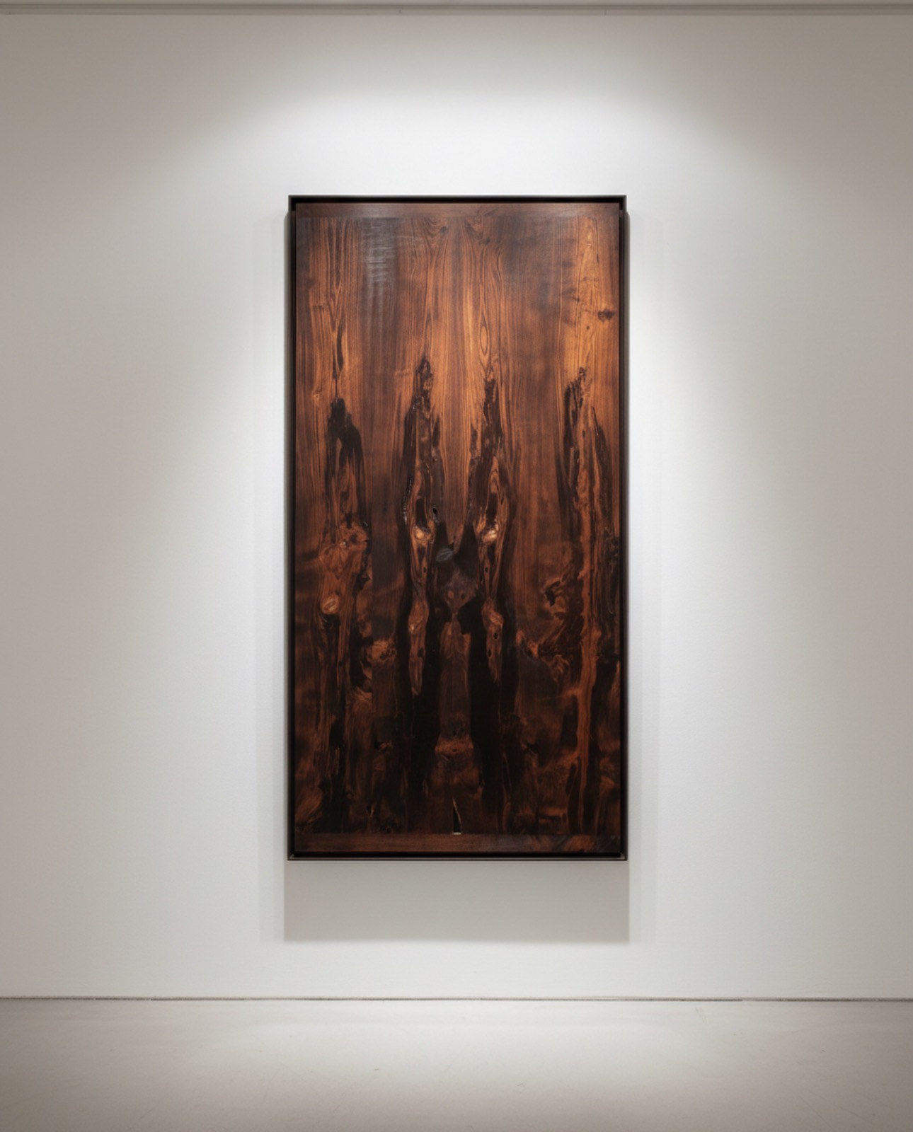 Vertical wooden artwork with dark and reddish tones hanging on a white wall, illuminated by overhead lighting.