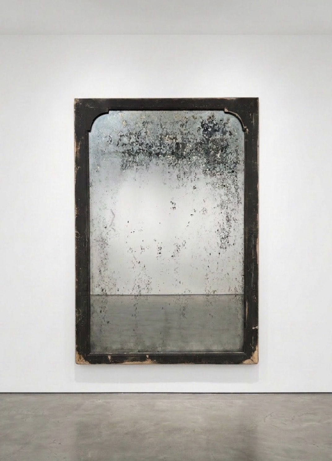 A dirty, weathered mirror with a black frame, mounted on a white wall, reflecting a plain surface.