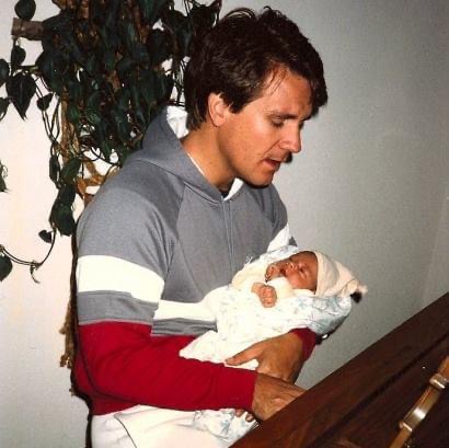 Young man holding a newborn baby wrapped in a blanket, looking down at the baby, standing near a wall with a plant.