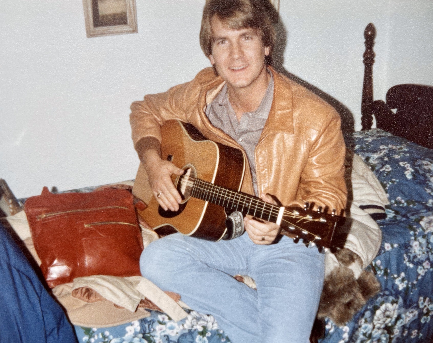 Young man sitting on a bed playing an acoustic guitar, wearing a tan leather jacket and light blue jeans.