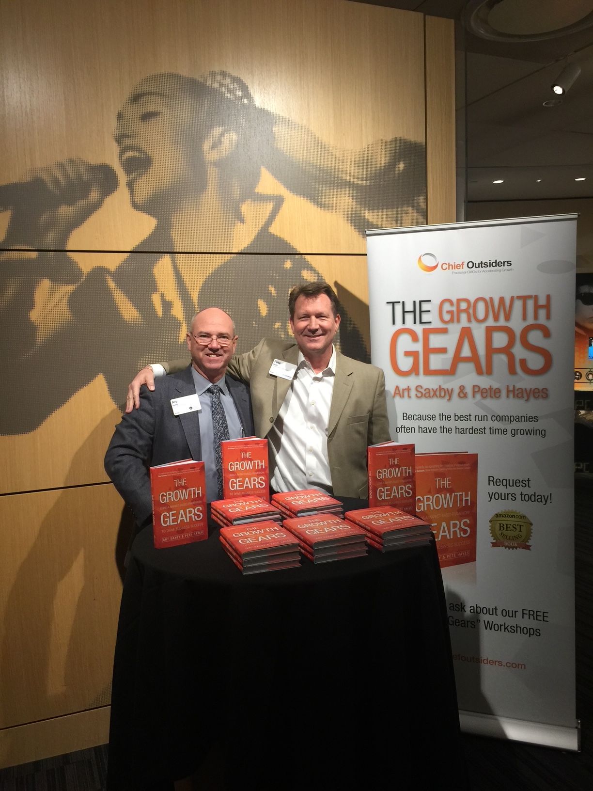 Two men standing behind a table with awards, in front of a banner about growth and geese, in a conference room.