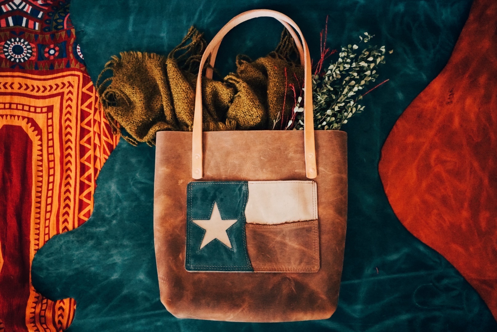 Brown leather tote bag with Texas flag patch, surrounded by colorful fabrics on a dark background.