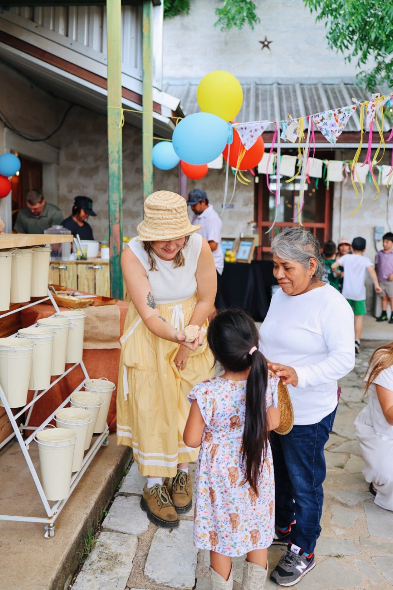 People at an outdoor celebration with balloons and decorations, including a woman in a hat and a girl talking to an older woman.