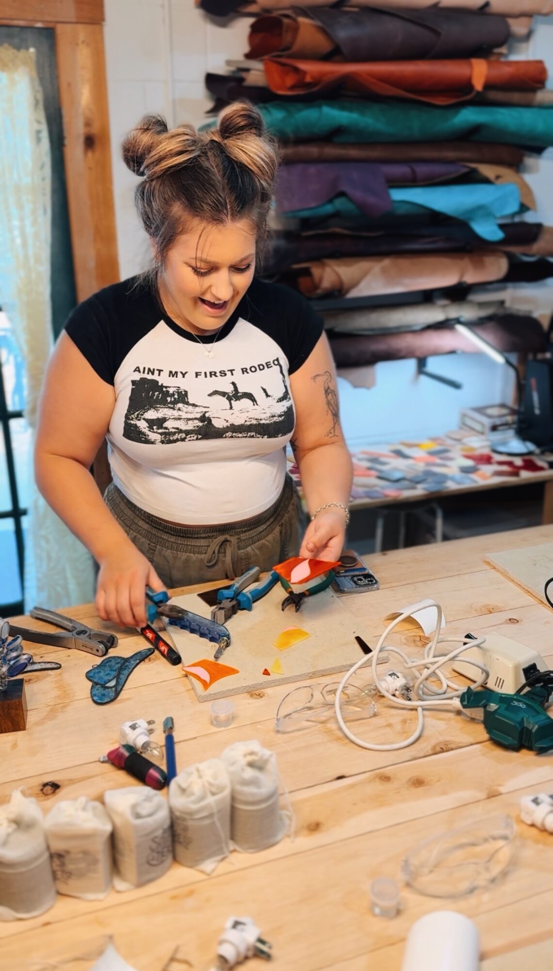 Young girl with two buns smiling at a worktable with fabric, tools, and spools of thread, surrounded by fabric stacks.