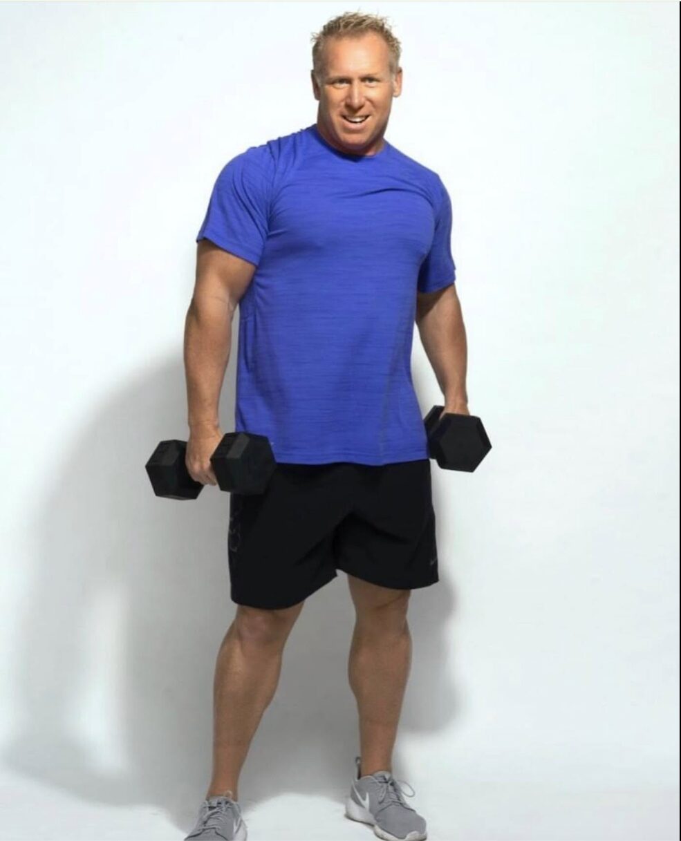 Man in blue shirt and black shorts holding dumbbells, smiling, standing against a white background.