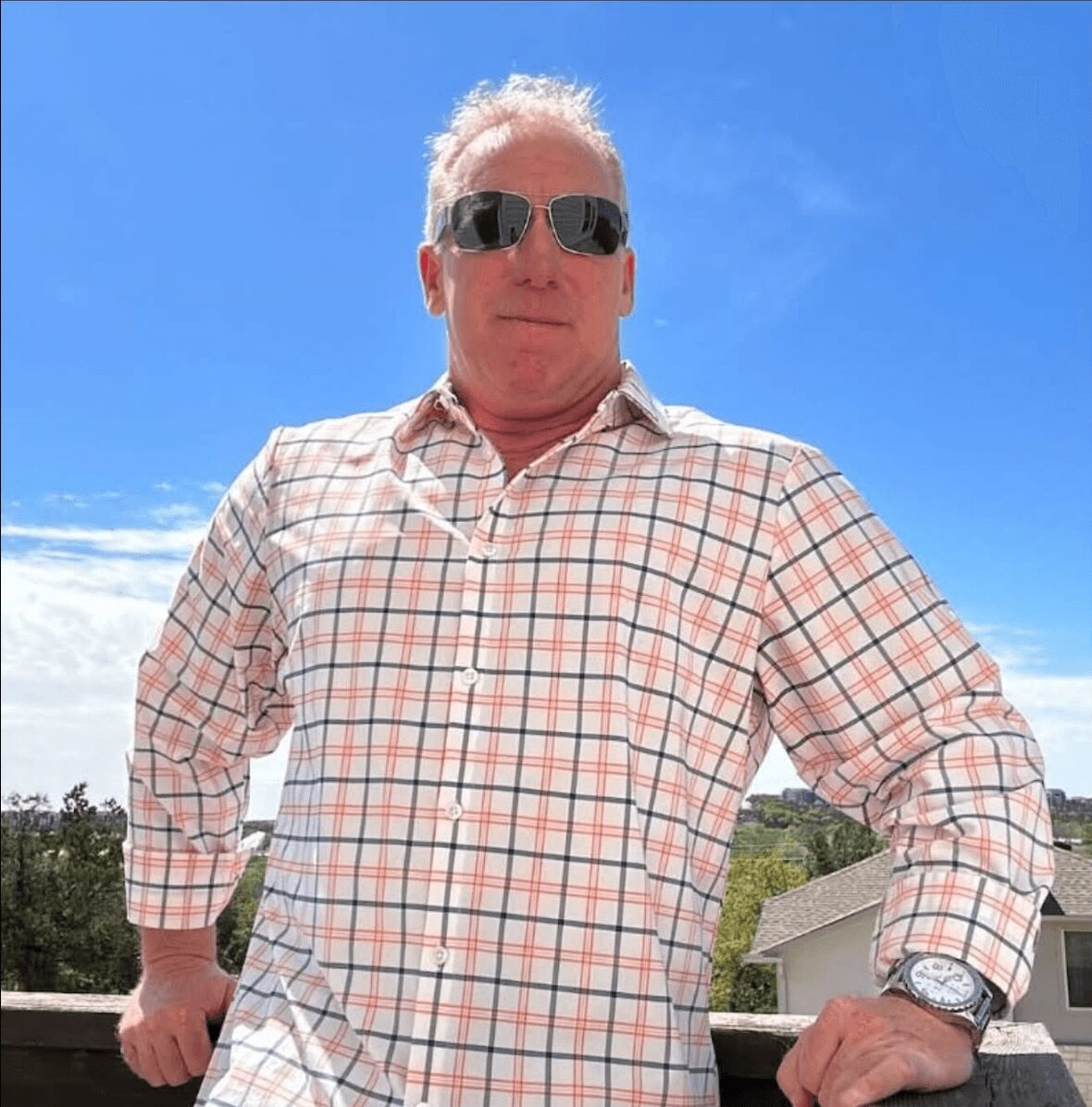Man wearing sunglasses and a checkered shirt outdoors on a sunny day, with a blue sky and trees in the background.