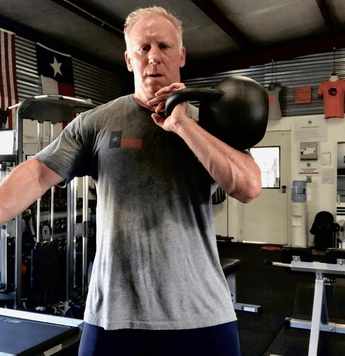 Man lifting a kettlebell in a gym, wearing a gray shirt and dark pants, with gym equipment in background.