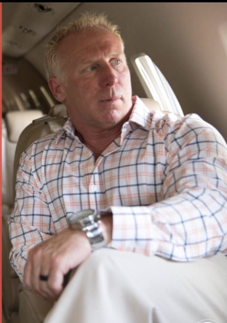 Man with blond hair wearing a checkered shirt, sitting in an airplane, looking out the window.