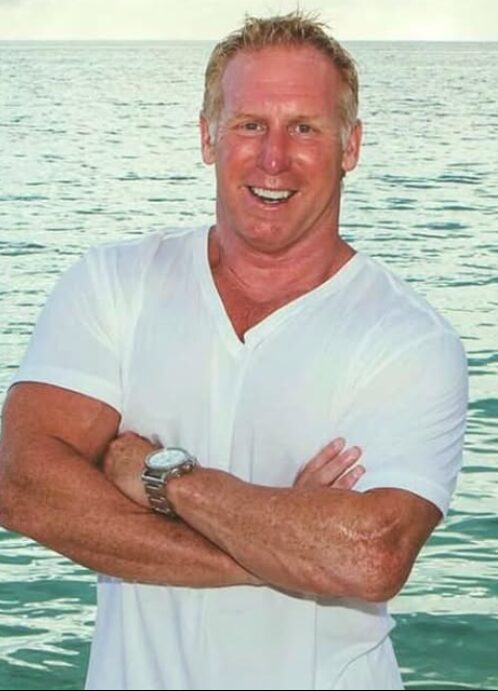 Man smiling with arms crossed, standing near water, wearing a white shirt, with a watch on his left wrist.