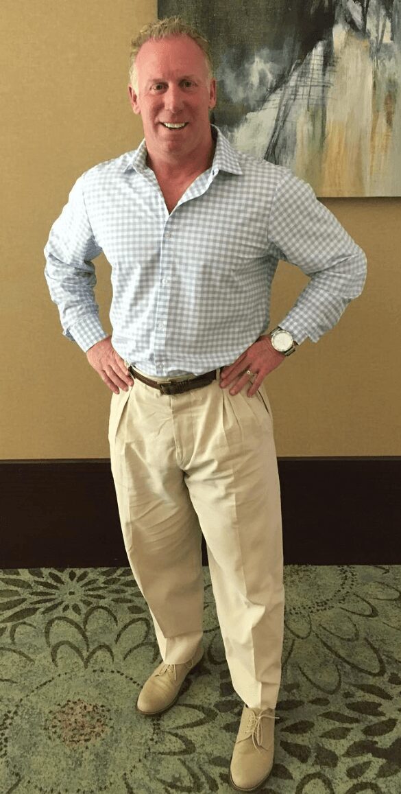 Man standing indoors with hands on hips, smiling, wearing a checkered shirt, beige pants, and a watch.