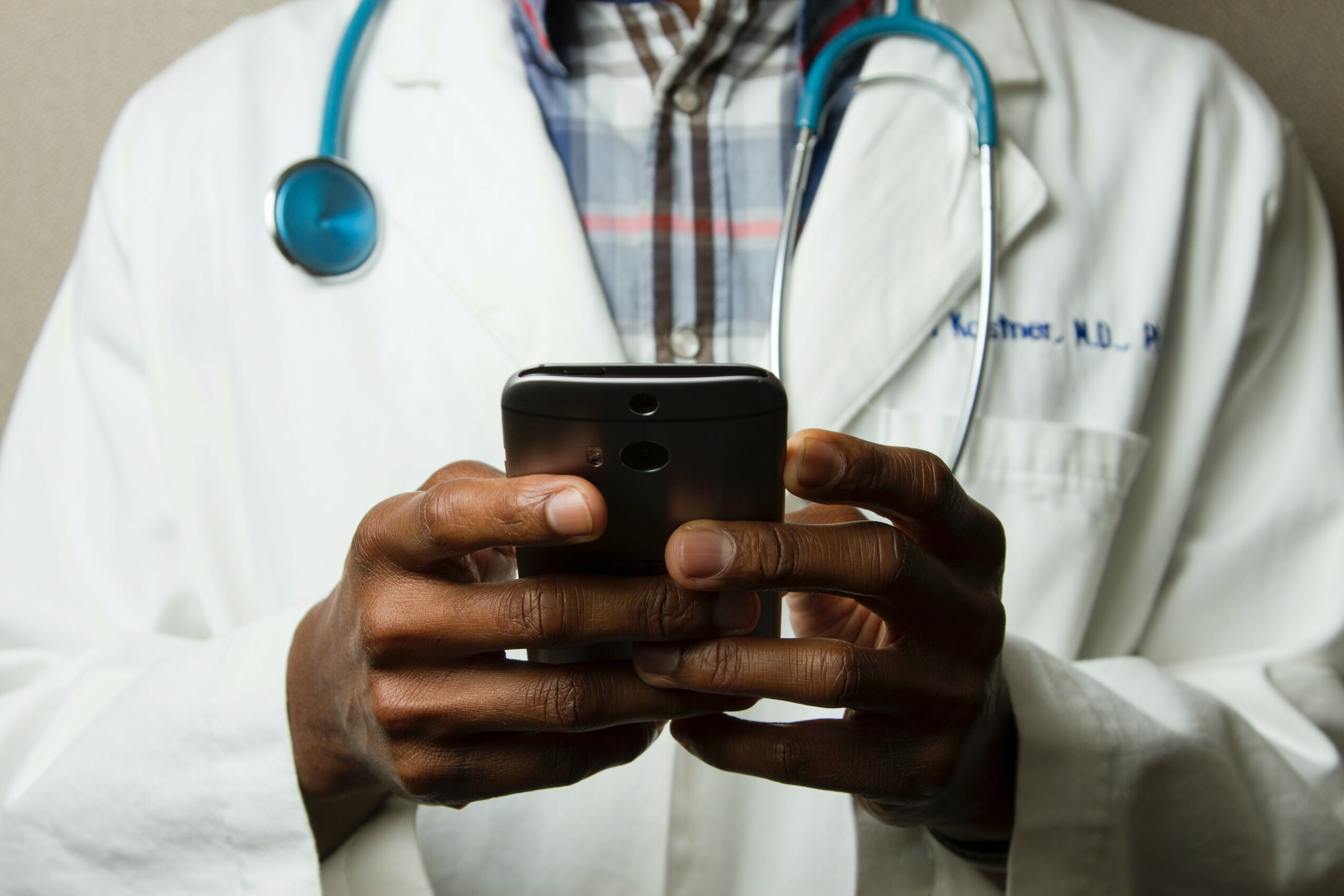Person in medical coat with stethoscope looking at smartphone, cropped at shoulders, neutral background.